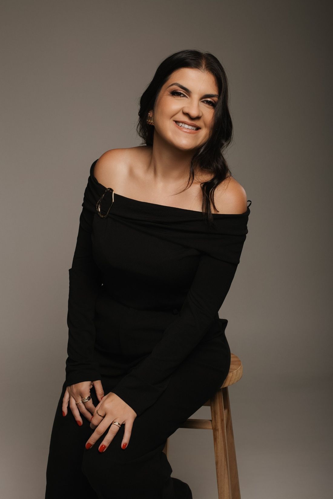 Fernanda sorrindo, sentada num banco de madeira, de blusa preta ombro a ombro